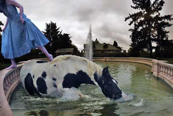 Horse pushes his nose through the water while walking in a fountain and a girl in a blue dress steps off his back
