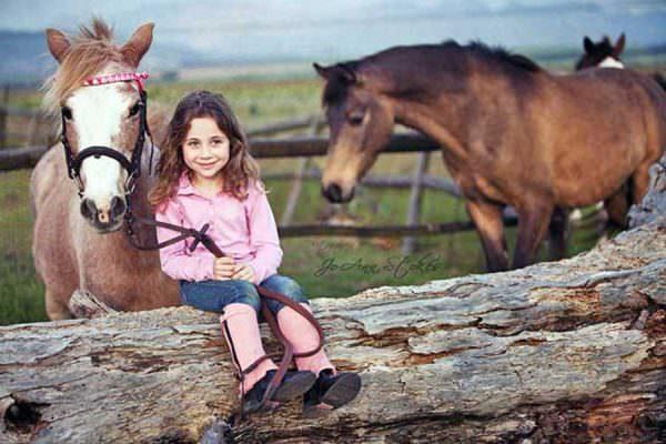 Young girl holding the reins of her roan pony while sitting on a log dressed in pink