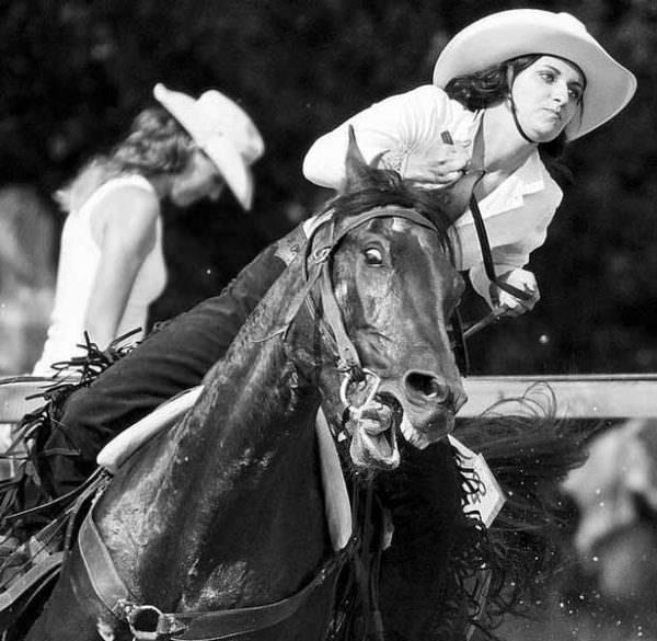A cowgirl pulls hard on her horse's mouth while the horse is wide-eyed and open mouthed in distress.