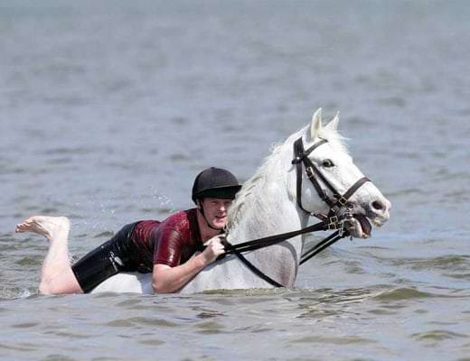 Prince Henry swimming with a horse and puliling on the horse's mouth.